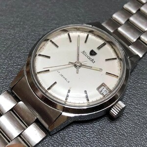Puede incluir: Un reloj de pulsera Nivada vintage con una correa de metal plateado y una esfera blanca. La esfera del reloj presenta marcadores de hora negros, el nombre de la marca y el texto "17 JEWELS". La fecha se muestra a las 3 en punto.