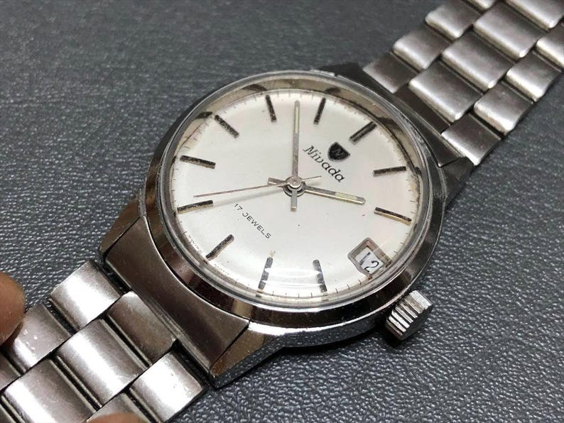 Puede incluir: Un reloj de pulsera Nivada vintage con una correa de metal plateado y una esfera blanca. La esfera del reloj presenta marcadores de hora negros, el nombre de la marca y el texto "17 JEWELS". La fecha se muestra a las 3 en punto.