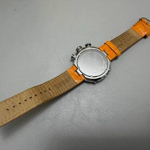 以下が含まれることがあります： オレンジ色の革バンドが付いたシルバーの腕時計。時計の文字盤は見えませんが、時計の裏側とバンドの裏側が見えます。バンドの裏側は薄茶色です。