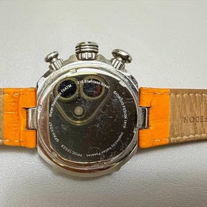 以下が含まれることがあります： オレンジ色の革ストラップが付いた、シルバーカラーのGiorgio Fedonの腕時計。 裏面には「Water Resistant 10ATM」や「316 Stainless Steel」などの文字が刻まれています。 ストラップは本革製です。