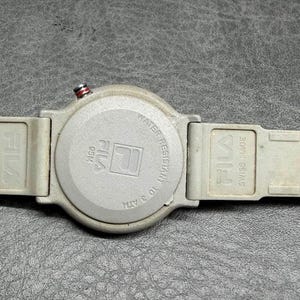 May include: A gray Fila watch with a round face and a gray band. The watch face has the Fila logo and the words "Water Resistant 10 ATM". The band is marked "Swiss Made".