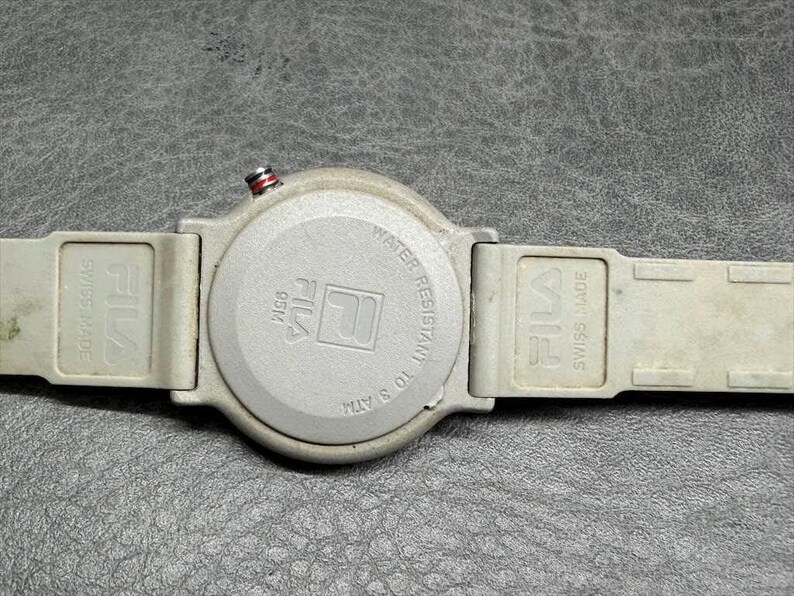 May include: A gray Fila watch with a round face and a gray band. The watch face has the Fila logo and the words "Water Resistant 10 ATM". The band is marked "Swiss Made".