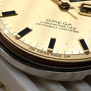 Puede incluir: Primer plano de una esfera de reloj Omega Chronometer dorada. El reloj presenta marcadores de hora y agujas rectangulares negros, con el d&iacute;a mostrado en una peque&ntilde;a ventana. El texto "OMEGA CHRONOMETER OFFICIALLY CERTIFIED" es visible.