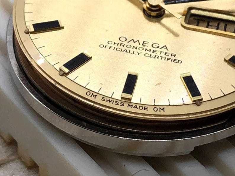 Puede incluir: Primer plano de una esfera de reloj Omega Chronometer dorada. El reloj presenta marcadores de hora y agujas rectangulares negros, con el d&iacute;a mostrado en una peque&ntilde;a ventana. El texto "OMEGA CHRONOMETER OFFICIALLY CERTIFIED" es visible.