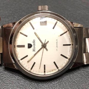 Puede incluir: Un reloj de pulsera Nivada con una correa y caja de metal plateado. La esfera del reloj es blanca con marcadores y agujas negras. El reloj tiene una ventana de fecha a las 3 en punto y el texto "17 JEWELS" y "SWISS MADE".