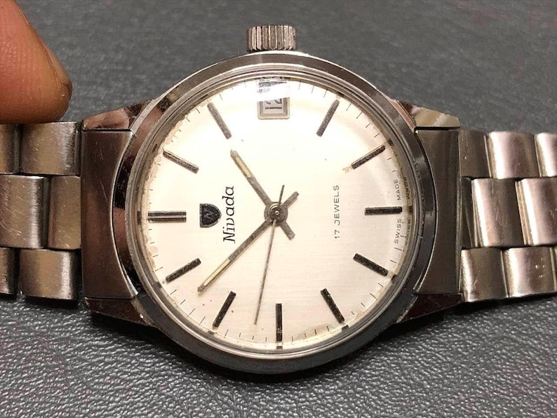 Puede incluir: Un reloj de pulsera Nivada con una correa y caja de metal plateado. La esfera del reloj es blanca con marcadores y agujas negras. El reloj tiene una ventana de fecha a las 3 en punto y el texto "17 JEWELS" y "SWISS MADE".