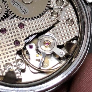 Puede incluir: Primer plano del mecanismo de un reloj con intrincados engranajes, tornillos y joyas. Los componentes plateados est&aacute;n dispuestos dentro de un marco circular, mostrando la precisi&oacute;n del funcionamiento interno del reloj. La palabra "Nevada" es visible.