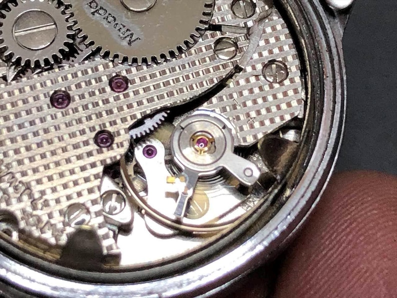 Puede incluir: Primer plano del mecanismo de un reloj con intrincados engranajes, tornillos y joyas. Los componentes plateados est&aacute;n dispuestos dentro de un marco circular, mostrando la precisi&oacute;n del funcionamiento interno del reloj. La palabra "Nevada" es visible.