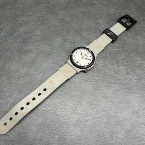 May include: A vintage FILA wristwatch with a white face, black bezel, and light gray band. The watch features black hour markers, red second hand, and the FILA logo. The band has multiple adjustment holes and a black buckle.
