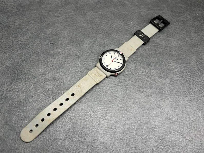 May include: A vintage FILA wristwatch with a white face, black bezel, and light gray band. The watch features black hour markers, red second hand, and the FILA logo. The band has multiple adjustment holes and a black buckle.