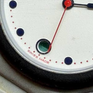 May include: Close-up of a wristwatch face with a white background, black bezel, and silver accents. The watch features a red second hand, black and blue hands, and blue hour markers. The text "SWISS MADE" is visible.