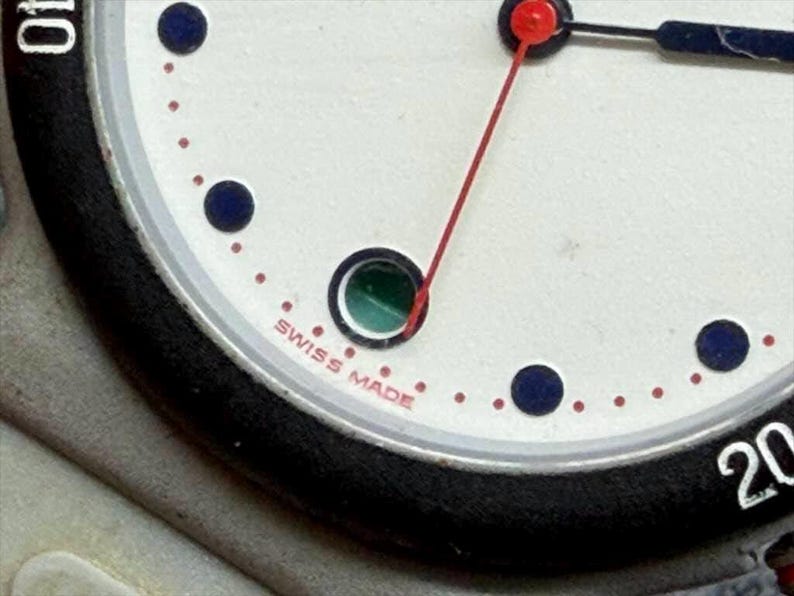 May include: Close-up of a wristwatch face with a white background, black bezel, and silver accents. The watch features a red second hand, black and blue hands, and blue hour markers. The text "SWISS MADE" is visible.
