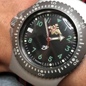 May include: A silver-toned wristwatch with a black face, featuring green numerals and a gold emblem. The watch has a rotating bezel with white markings and a date display. The word "РОССИЯ" is visible on the watch face.