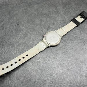 May include: A FILA brand wristwatch with a light gray band and a black buckle. The watch face is silver with a gray band. The watch is lying on a dark gray surface.