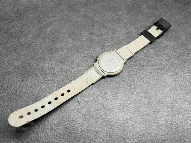May include: A FILA brand wristwatch with a light gray band and a black buckle. The watch face is silver with a gray band. The watch is lying on a dark gray surface.