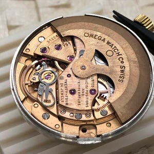 Puede incluir: Primer plano del movimiento de un reloj Omega, que muestra intrincados engranajes, joyas y la inscripci&oacute;n "OMEGA WATCH CO SWISS". El movimiento es un complejo conjunto de componentes dorados y plateados, que destaca la precisi&oacute;n y la artesan&iacute;a.