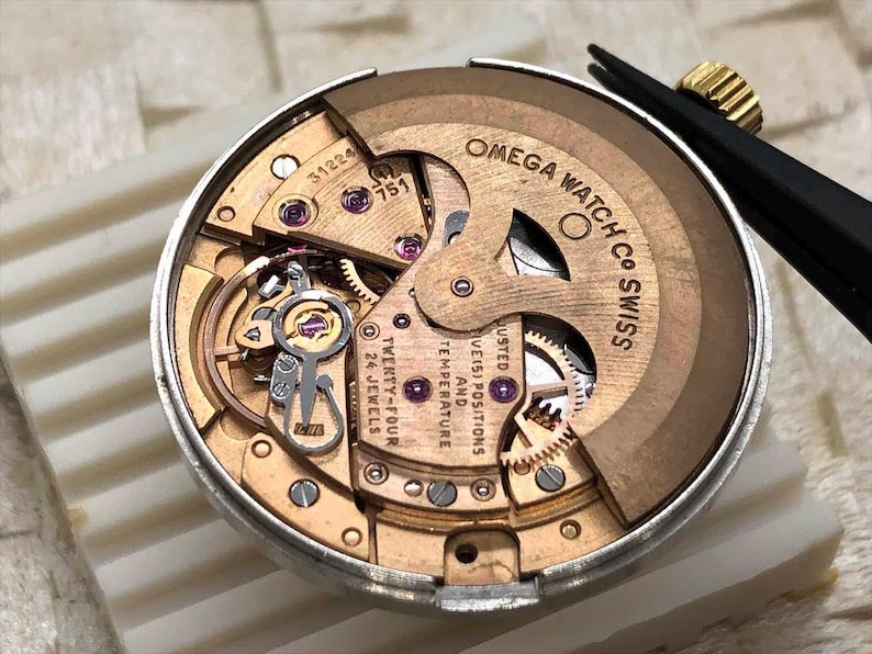 Puede incluir: Primer plano del movimiento de un reloj Omega, que muestra intrincados engranajes, joyas y la inscripci&oacute;n "OMEGA WATCH CO SWISS". El movimiento es un complejo conjunto de componentes dorados y plateados, que destaca la precisi&oacute;n y la artesan&iacute;a.