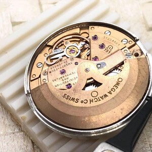 Puede incluir: Primer plano del movimiento de un reloj Omega, con tonos dorados, plateados y cobrizos. El mecanismo del reloj est&aacute; parcialmente desmontado, revelando engranajes y componentes intrincados. El texto "OMEGA WATCH CO SWISS" es visible.