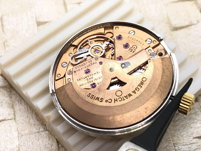 Puede incluir: Primer plano del movimiento de un reloj Omega, con tonos dorados, plateados y cobrizos. El mecanismo del reloj est&aacute; parcialmente desmontado, revelando engranajes y componentes intrincados. El texto "OMEGA WATCH CO SWISS" es visible.