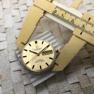 Puede incluir: Una esfera de reloj autom&aacute;tico Omega Constellation dorada con marcadores y agujas negras, y una pantalla de fecha. Un calibrador beige est&aacute; midiendo la esfera del reloj. La esfera del reloj est&aacute; sobre una superficie blanca y gris.
