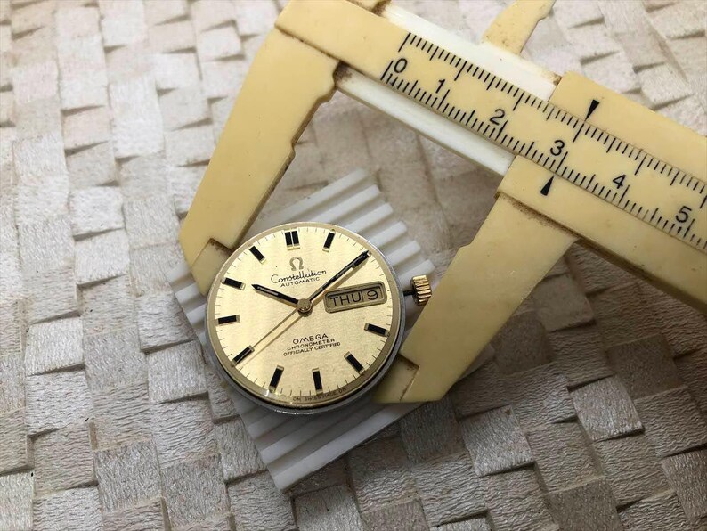 Puede incluir: Una esfera de reloj autom&aacute;tico Omega Constellation dorada con marcadores y agujas negras, y una pantalla de fecha. Un calibrador beige est&aacute; midiendo la esfera del reloj. La esfera del reloj est&aacute; sobre una superficie blanca y gris.