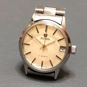 Puede incluir: Un reloj de pulsera Nivada vintage con caja y correa plateadas. La esfera crema presenta marcadores de hora negros, agujas doradas y una ventana de fecha. Est&aacute; marcado como "17 JEWELS" y "SWISS MADE". La caja mide aproximadamente 3,5 cm de di&aacute;metro.