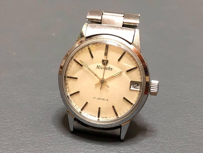 Puede incluir: Un reloj de pulsera Nivada vintage con caja y correa plateadas. La esfera crema presenta marcadores de hora negros, agujas doradas y una ventana de fecha. Est&aacute; marcado como "17 JEWELS" y "SWISS MADE". La caja mide aproximadamente 3,5 cm de di&aacute;metro.