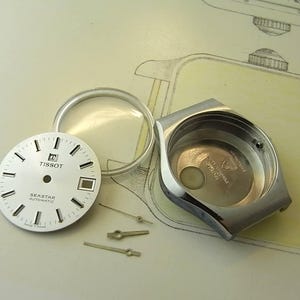 May include: Disassembled Tissot Seastar Automatic watch components. Includes a silver watch case, a white watch face with black markers and hands, a clear crystal, and small silver watch hands. The watch face reads "Tissot Seastar Automatic".
