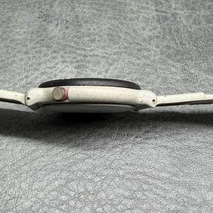 May include: Side view of a vintage white wristwatch with a black watch face and a red button. The watch has a white band and is set against a textured gray surface.