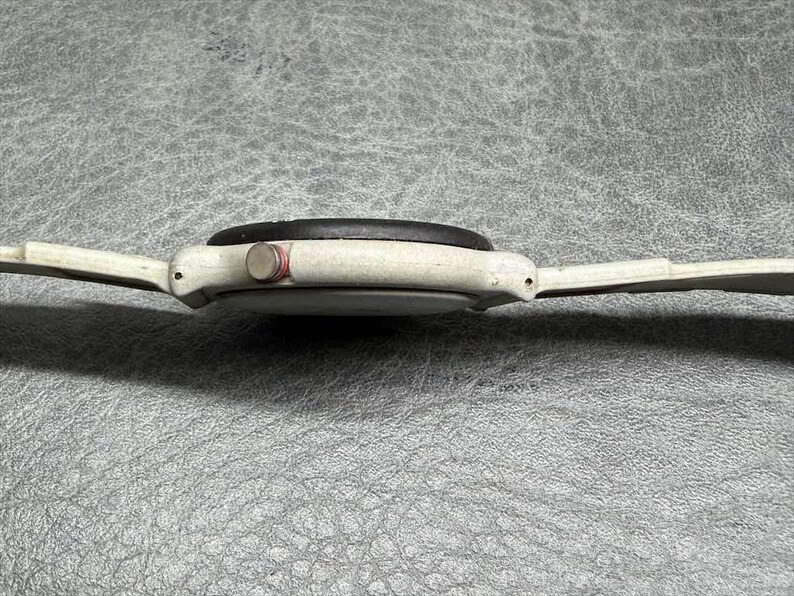 May include: Side view of a vintage white wristwatch with a black watch face and a red button. The watch has a white band and is set against a textured gray surface.