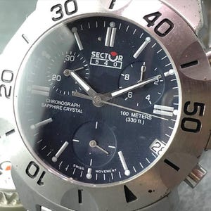 May include: A silver-toned Sector 540 chronograph watch with a dark blue face. The watch features a sapphire crystal, multiple sub-dials, and is water-resistant to 100 meters (330 ft). The bezel has prominent numerals.