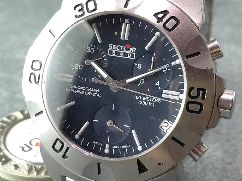 May include: A silver-toned Sector 540 chronograph watch with a dark blue face. The watch features a sapphire crystal, multiple sub-dials, and is water-resistant to 100 meters (330 ft). The bezel has prominent numerals.