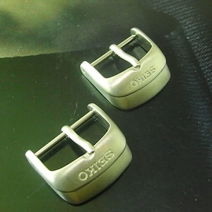 May include: Two silver-toned metal watch buckles with a brushed finish. Each buckle features a rectangular frame with a central prong and the brand name SEIKO engraved in capital letters on the front surface of the buckle base.