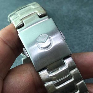 May include: Close-up of a silver-toned watch band clasp. The clasp features a stylized emblem and a secure locking mechanism. The watch band is made of interlocking metal links.