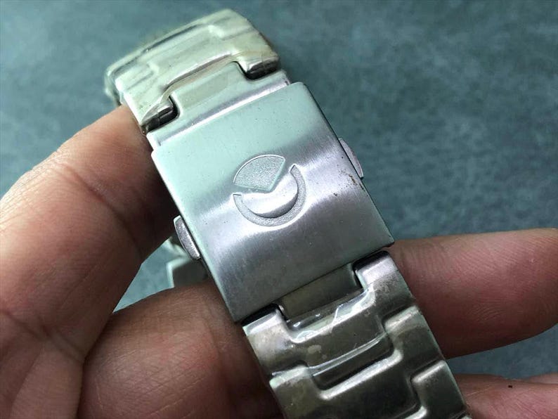 May include: Close-up of a silver-toned watch band clasp. The clasp features a stylized emblem and a secure locking mechanism. The watch band is made of interlocking metal links.