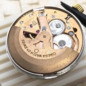 Puede incluir: Primer plano de un movimiento de reloj con intrincados engranajes y componentes. El mecanismo del reloj presenta el texto "OMEGA WATCH CO. SWISS" y otras marcas. Unas pinzas negras con punta dorada se encuentran en la esquina superior derecha.