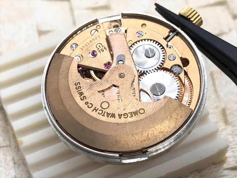 Puede incluir: Primer plano de un movimiento de reloj con intrincados engranajes y componentes. El mecanismo del reloj presenta el texto "OMEGA WATCH CO. SWISS" y otras marcas. Unas pinzas negras con punta dorada se encuentran en la esquina superior derecha.