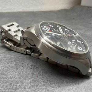 May include: A stainless steel wristwatch with a black dial, featuring white Arabic numerals, a date window, and the text "KRAFT" and "GMT" printed on the face. The watch has a brushed metal link bracelet with a folding clasp mechanism.