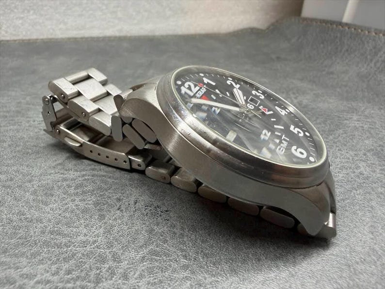 May include: A stainless steel wristwatch with a black dial, featuring white Arabic numerals, a date window, and the text "KRAFT" and "GMT" printed on the face. The watch has a brushed metal link bracelet with a folding clasp mechanism.