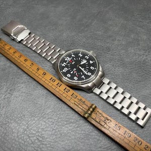 May include: A silver-toned stainless steel wristwatch with a black dial featuring white Arabic numerals, a date window, and a red-tipped GMT hand. The watch is positioned next to a wooden folding ruler showing a length of approximately 8.5 inches.