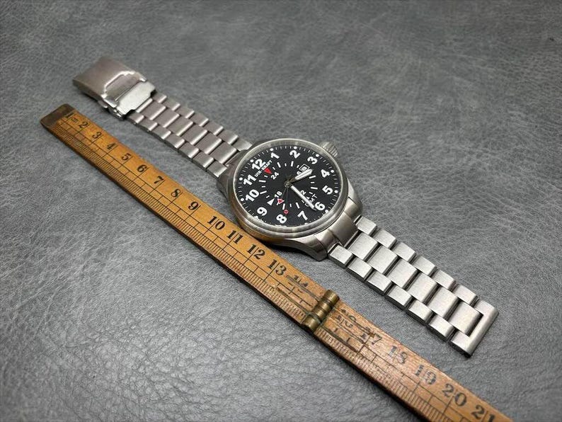 May include: A silver-toned stainless steel wristwatch with a black dial featuring white Arabic numerals, a date window, and a red-tipped GMT hand. The watch is positioned next to a wooden folding ruler showing a length of approximately 8.5 inches.
