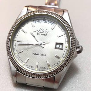 Puede incluir: Un reloj Mido Ocean Star de cuarzo plateado. La esfera es plateada clara con manecillas y marcadores negros, y una pantalla de fecha. El bisel es texturizado y la correa del reloj también es plateada.