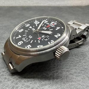 May include: A stainless steel wristwatch with a black dial featuring white Arabic numerals, a GMT function, and a date window. The watch has a brushed metal finish, a prominent crown on the side, and a matching metal link bracelet with a clasp.