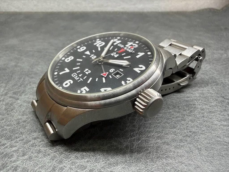 May include: A stainless steel wristwatch with a black dial featuring white Arabic numerals, a GMT function, and a date window. The watch has a brushed metal finish, a prominent crown on the side, and a matching metal link bracelet with a clasp.
