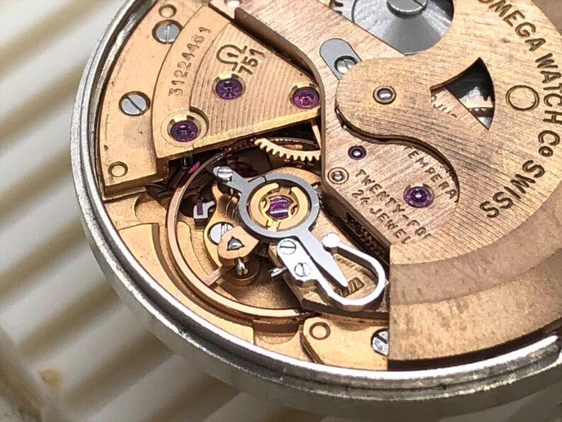 Puede incluir: Primer plano de un movimiento de reloj con intrincados engranajes y componentes. El mecanismo del reloj presenta placas doradas, rub&iacute;es y el texto "OMEGA WATCH CO SWISS". El reloj tiene el texto "TWENTY-FOUR 24 JEWEL".