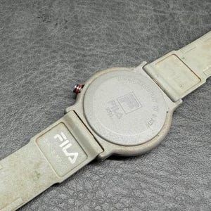 May include: A Fila watch with a light gray band and watch face. The watch face has the Fila logo and the words "Water Resistant to 5 ATM". The band is made of a flexible material.