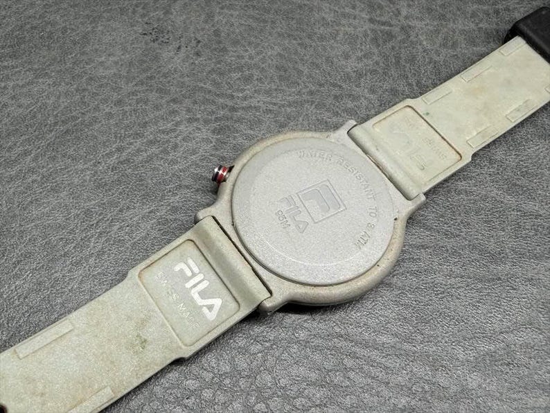 May include: A Fila watch with a light gray band and watch face. The watch face has the Fila logo and the words "Water Resistant to 5 ATM". The band is made of a flexible material.
