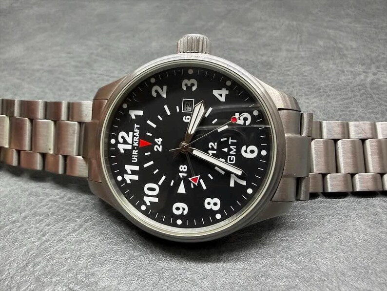 May include: A stainless steel wristwatch with a black dial, featuring white Arabic numerals, a date window, and a GMT function. The watch has a brushed metal link bracelet, a crown at the top, and the text UHR-KRAFT printed on the watch face.