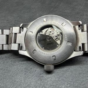 May include: A silver-toned stainless steel wristwatch with a metal link bracelet. The case back features a circular glass exhibition window revealing the internal mechanical movement, with text engraved around the perimeter reading ALL STAINLESS STEEL.