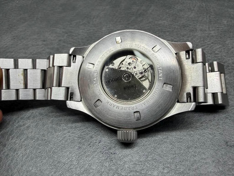 May include: A silver-toned stainless steel wristwatch with a metal link bracelet. The case back features a circular glass exhibition window revealing the internal mechanical movement, with text engraved around the perimeter reading ALL STAINLESS STEEL.